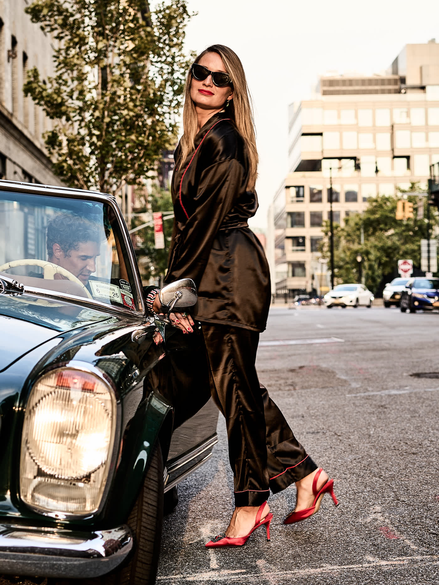 A model in an olive satin suit and red heels walking past the vintage green Mercedes on a Manhattan street