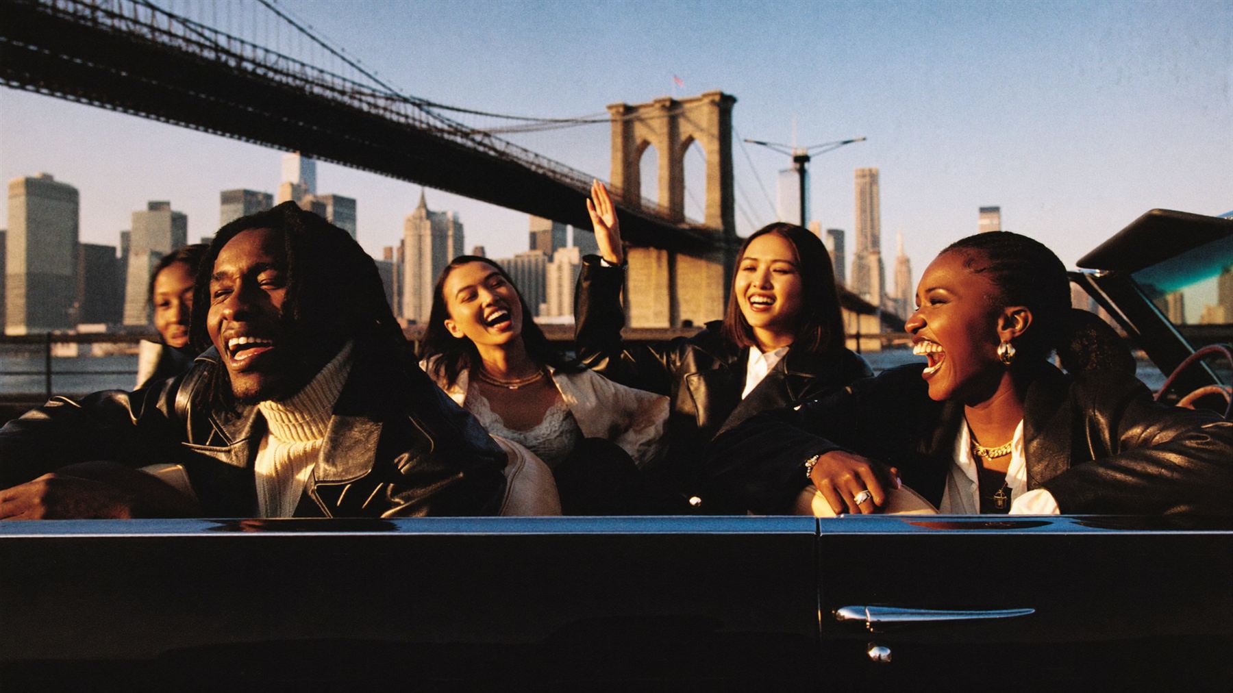 Five friends laughing in a vintage convertible under the Brooklyn Bridge at golden hour