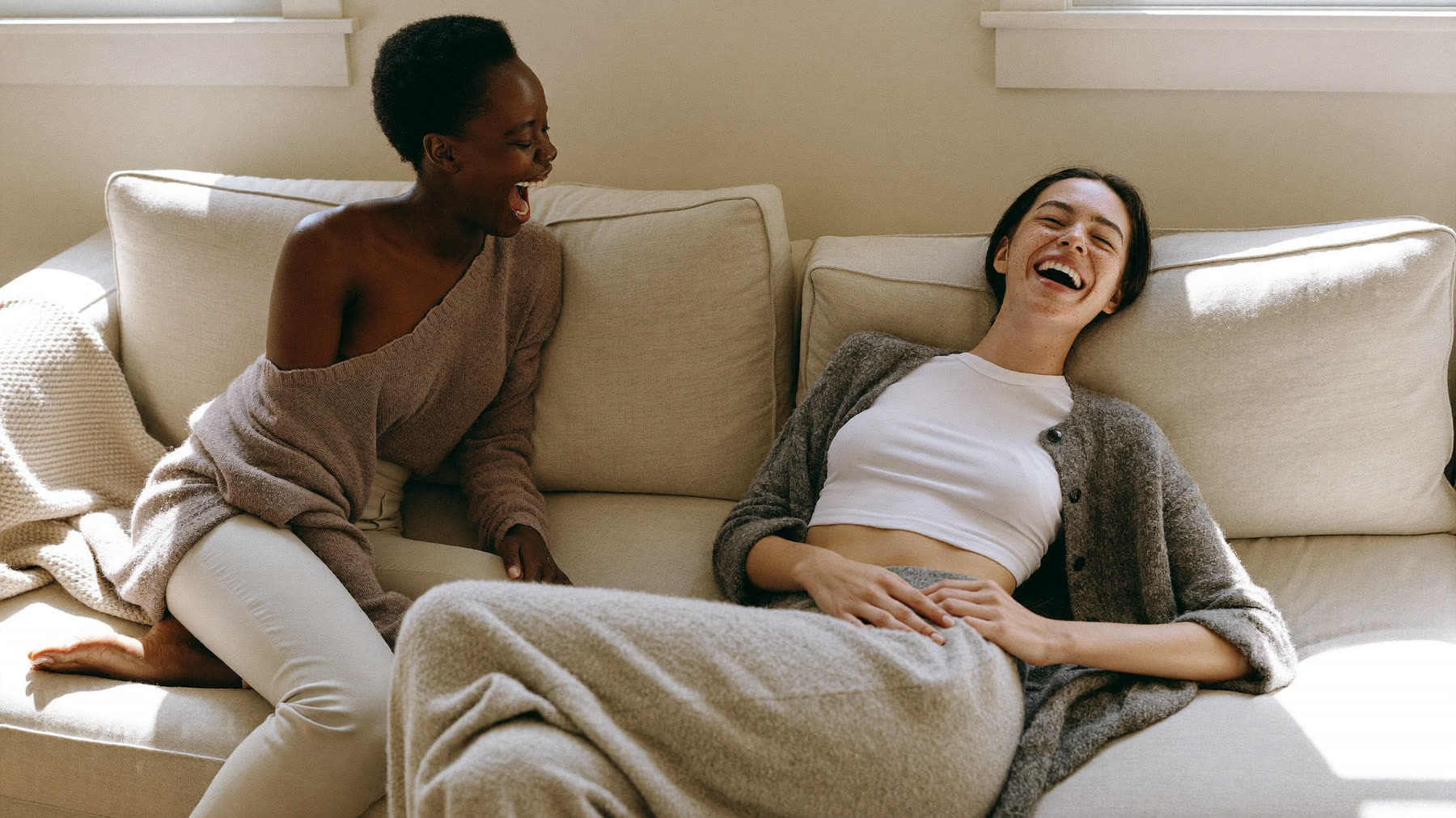 Two friends laughing together on a cream linen sofa in soft daylight