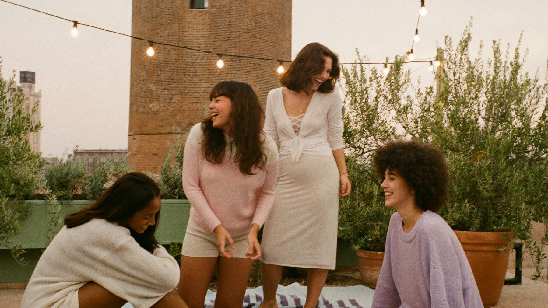 Four friends laughing on a Manhattan rooftop terrace under string lights at dusk